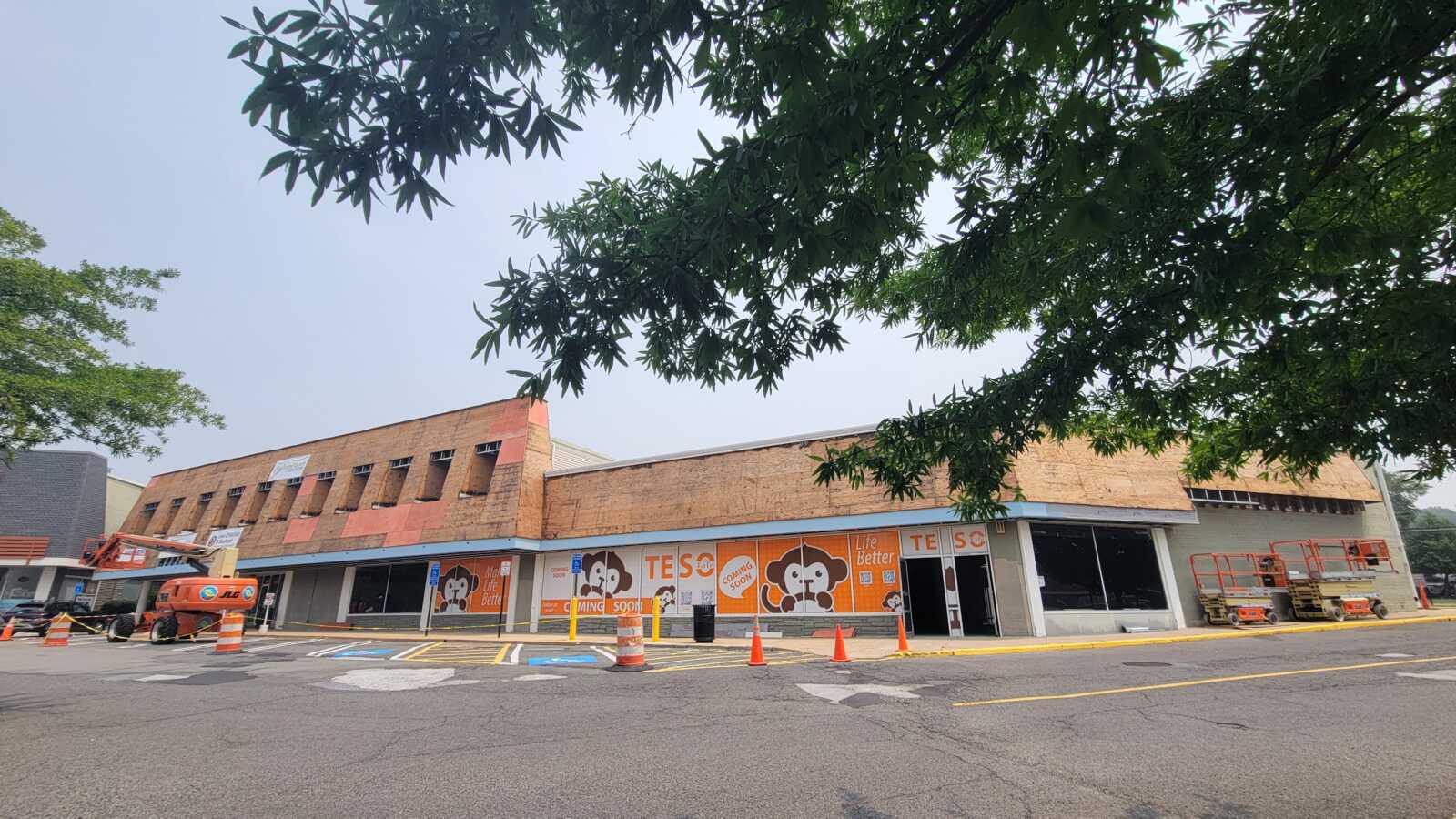 Shopfront under renovation with plywood siding, orange construction banners, and a cherry picker in the parking lot amid orange cones and barricades.
