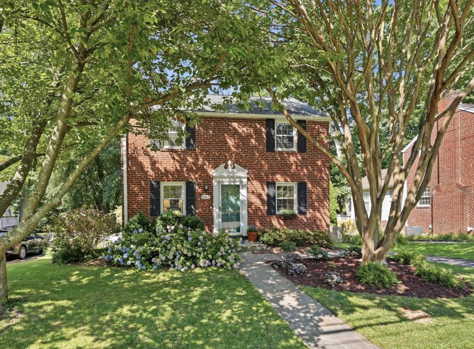 Brick two-story house with blue shutters, white front door, and a lush front yard shaded by large trees.