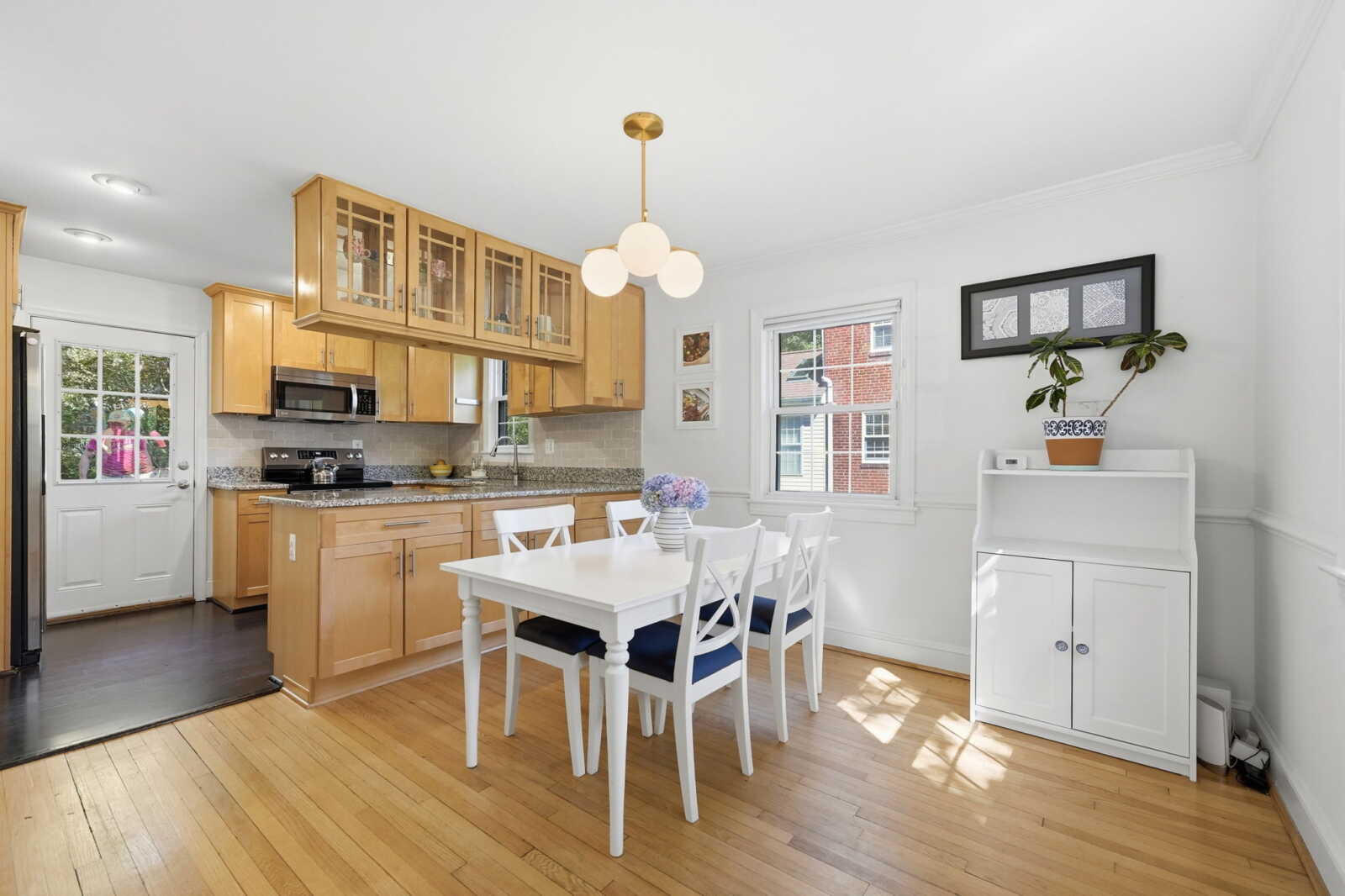 Bright open kitchen with wooden cabinets, a white dining table and chairs, and a white hutch by a window; potted plant on the shelf.
