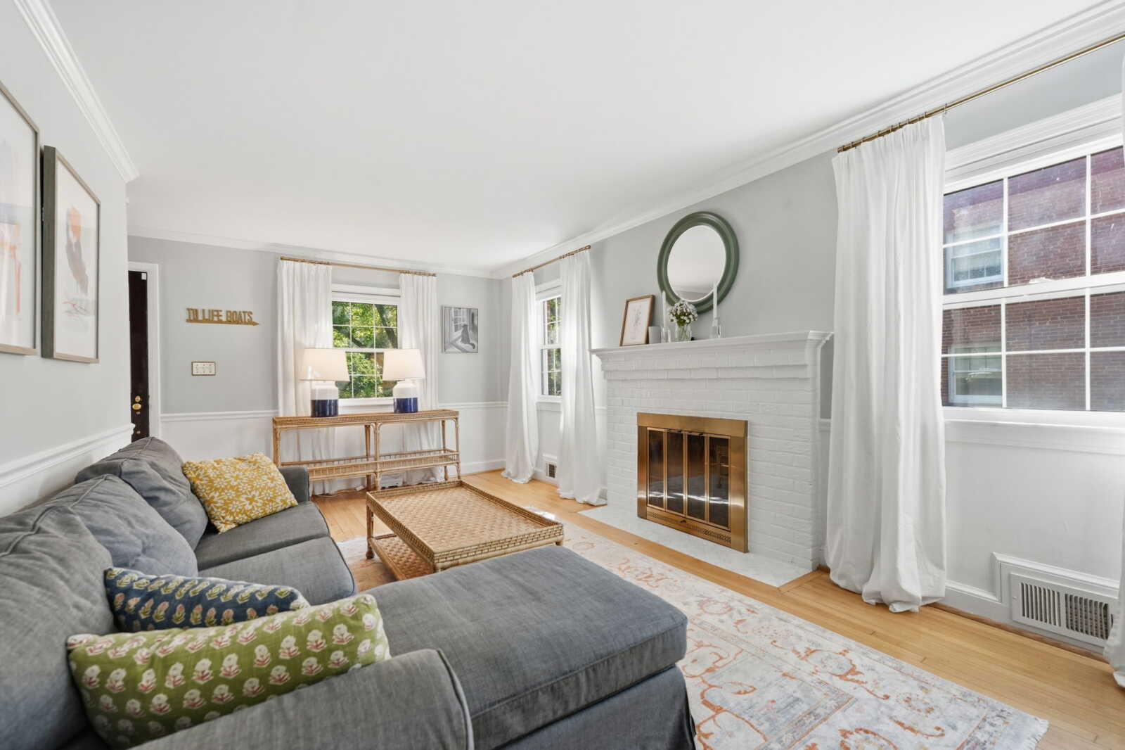 Bright living room with gray sectional, wicker coffee table, white brick fireplace, and large windows with white curtains.