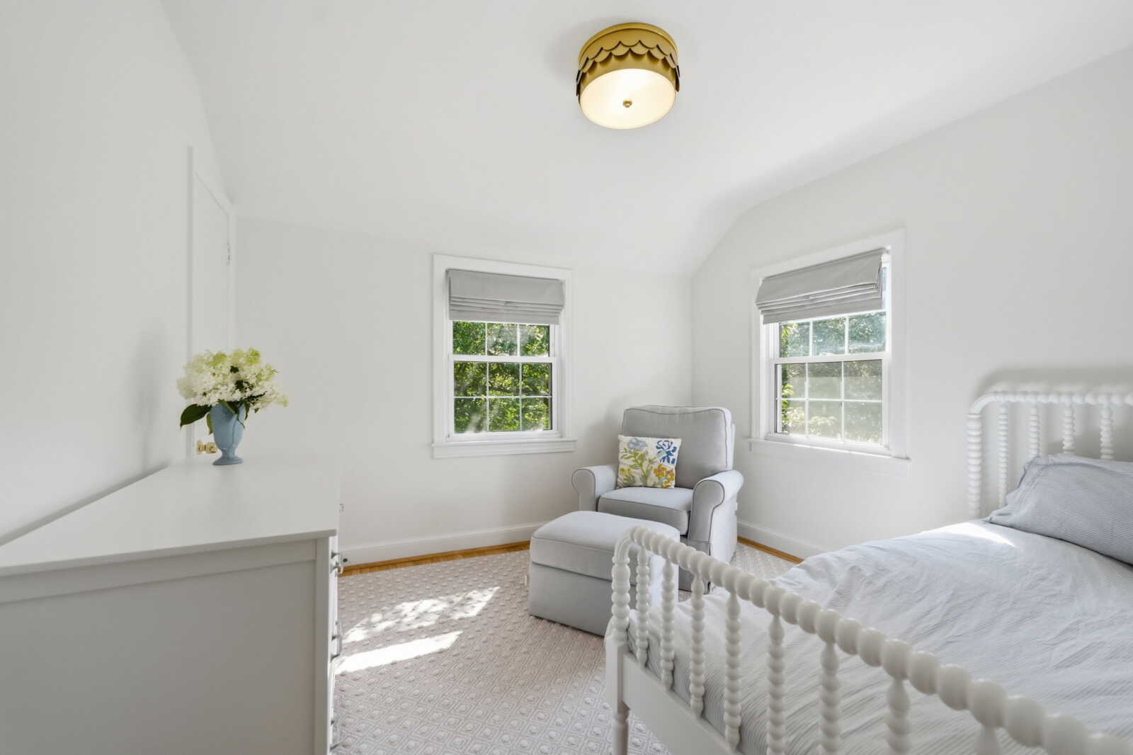 Bright white bedroom with a white spindle bed, gray armchair and ottoman, dresser with a vase of white flowers, and two windows.