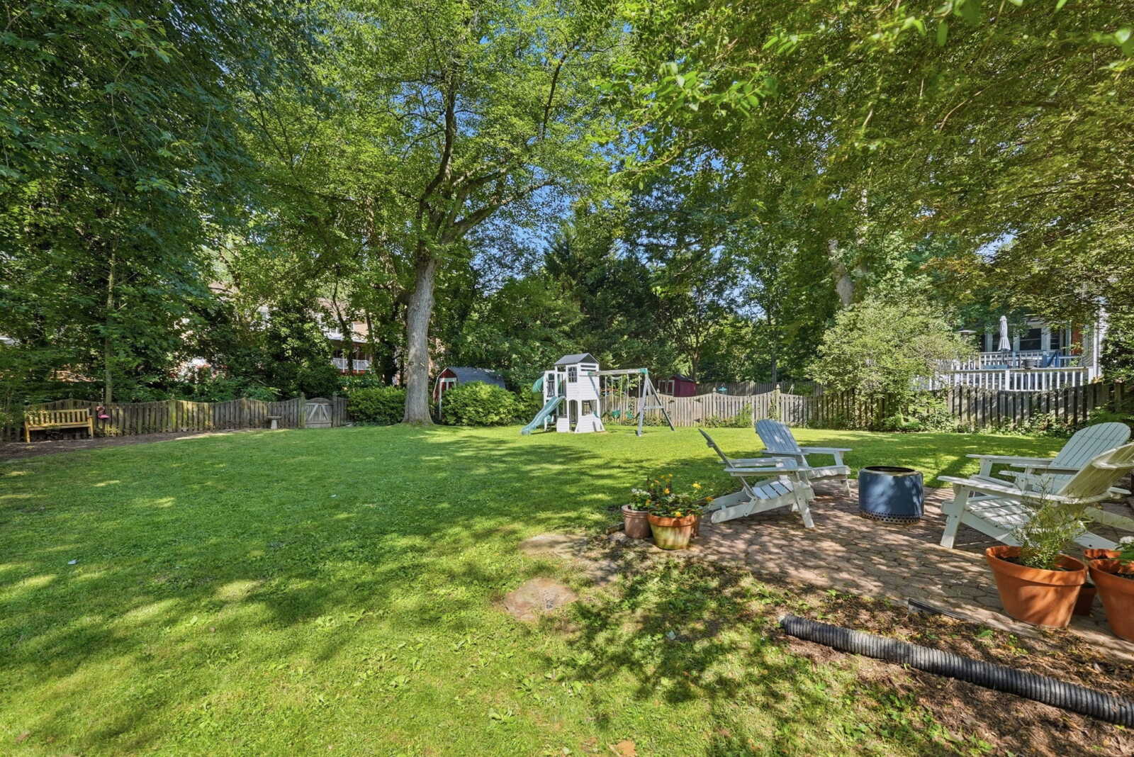 Spacious backyard with green lawn, trees, and a white playset featuring a slide in the center, surrounded by a wooden fence.