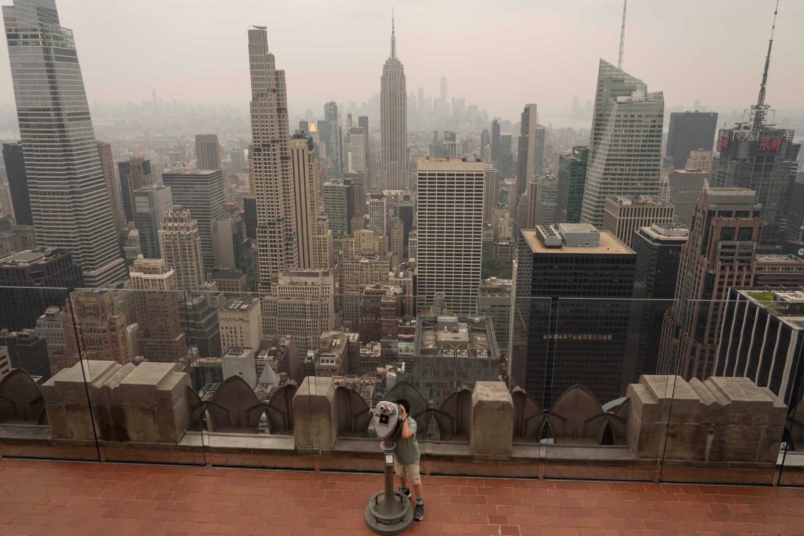 A person uses binoculars to look out as smoke from wildfires blankets the sky at Top of The Rock observation deck at the Rockefeller Center, Thursday, July 16, 2026, in New York. (AP Photo/Yuki Iwamura) | FFXnow Person using a public viewing telescope on an observation deck overlooking a dense, hazy city skyline.