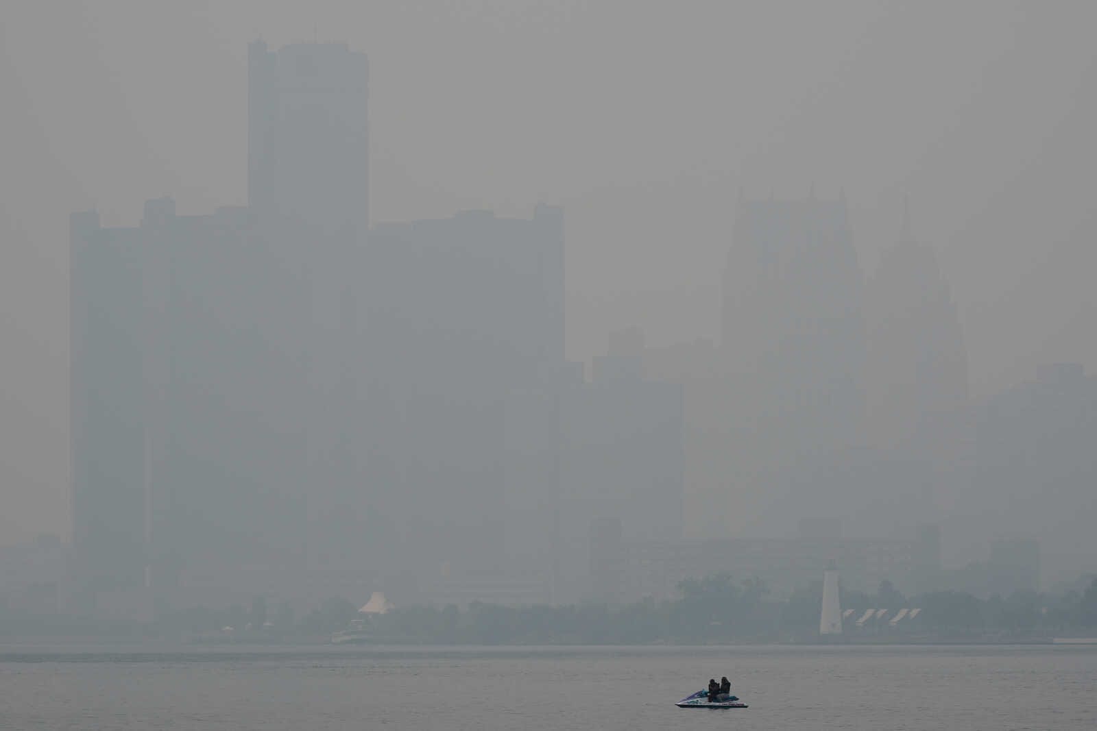 Hazy city skyline across a calm river; a small motorboat with two people in the foreground.