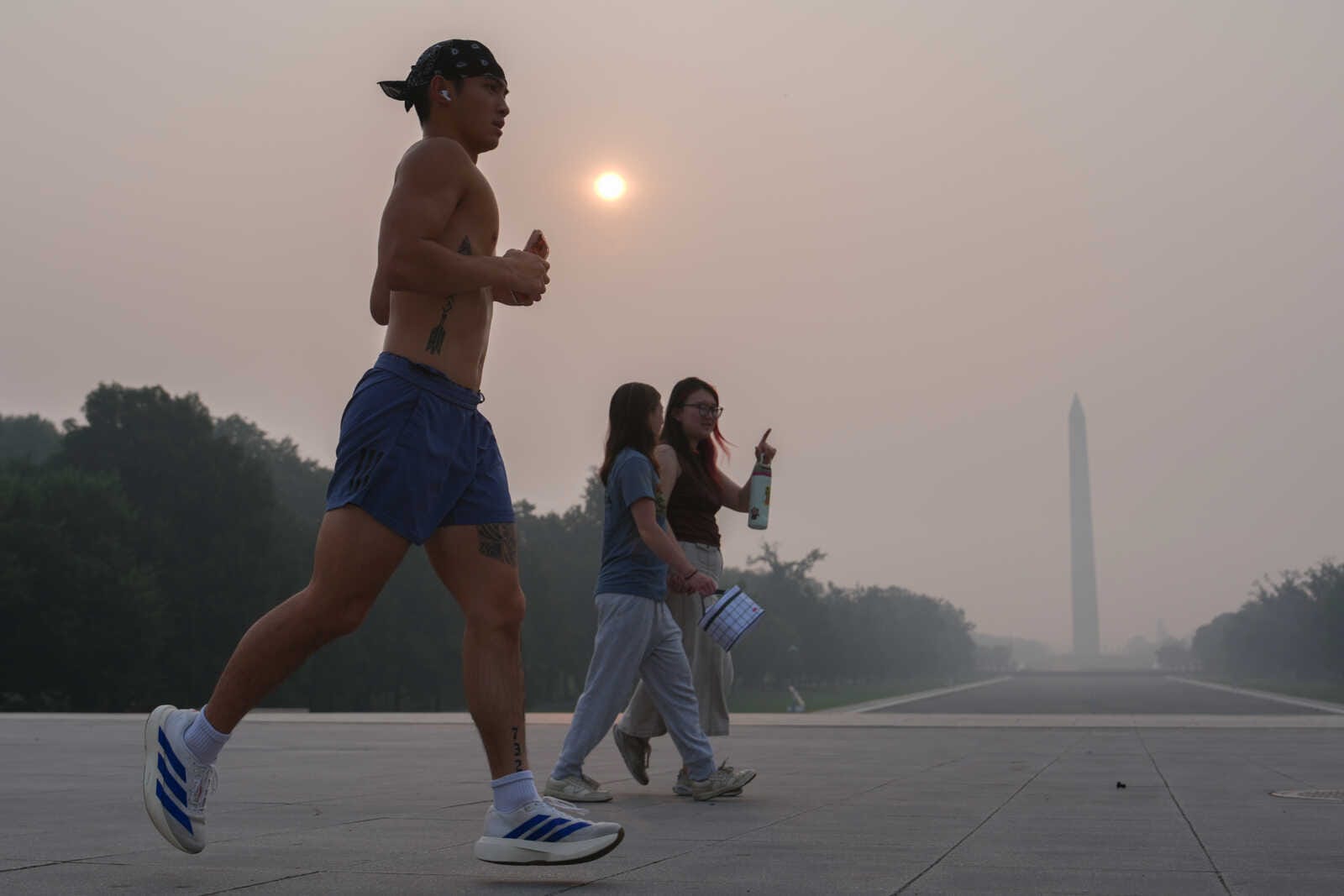 Shirtless man running in blue shorts on a wide plaza with two pedestrians and the Washington Monument in a hazy sunset background.