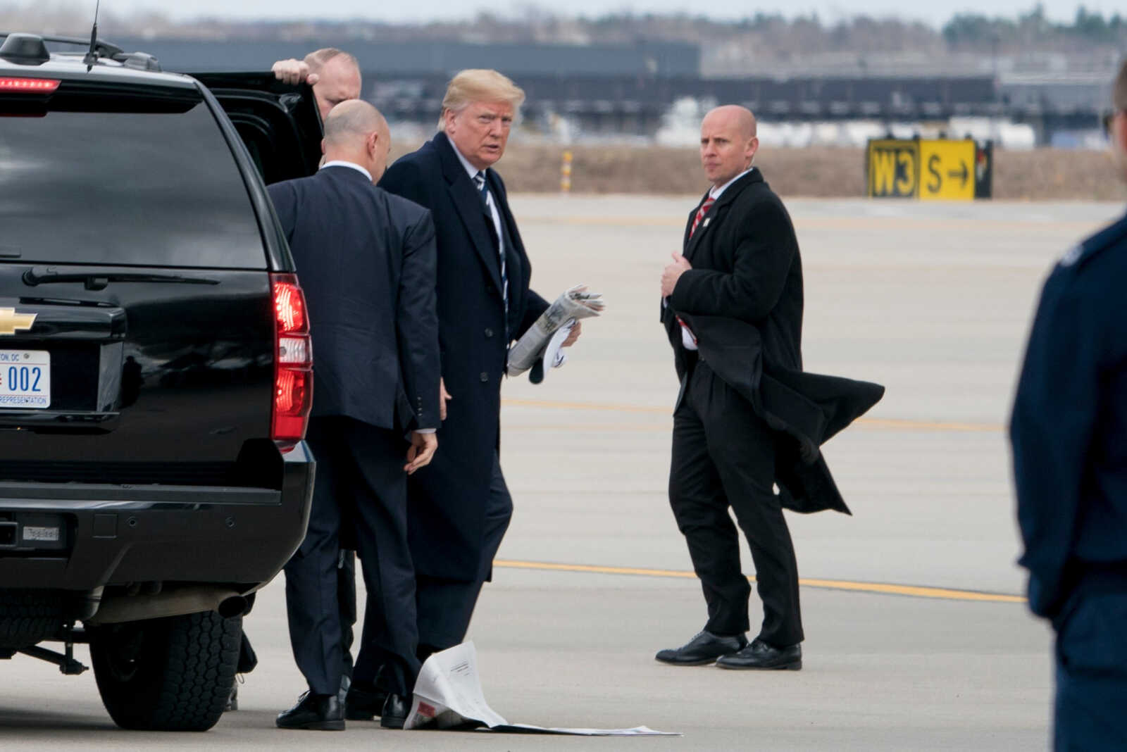 Former President Donald Trump stepping from a black SUV onto the airport tarmac, flanked by security and aides, with newspapers in hand.