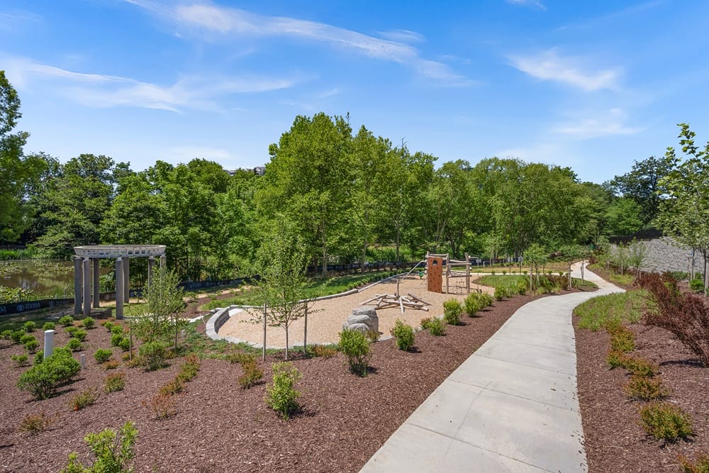 Sunny park scene with a curved concrete path, trees, and a wooden play structure in a mulch-covered play area near a river.