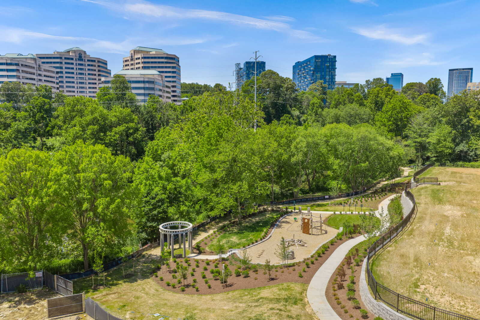 A lush urban park with a winding path, playground equipment, and a white circular gazebo, with high-rise buildings in the background under a blue sky.