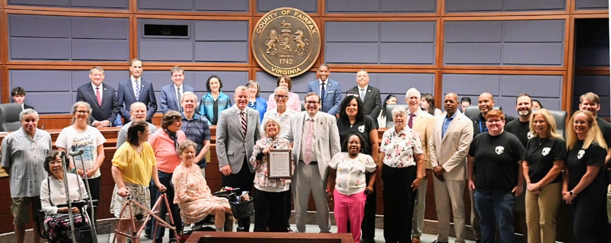 Bryna Helfer being honored by the Fairfax County Board of Supervisors (courtesy Fairfax County) | FFXnow Group of diverse adults in a formal meeting room, some in suits, posing with a certificate in front of a county seal that reads Virginia/Fairfax.