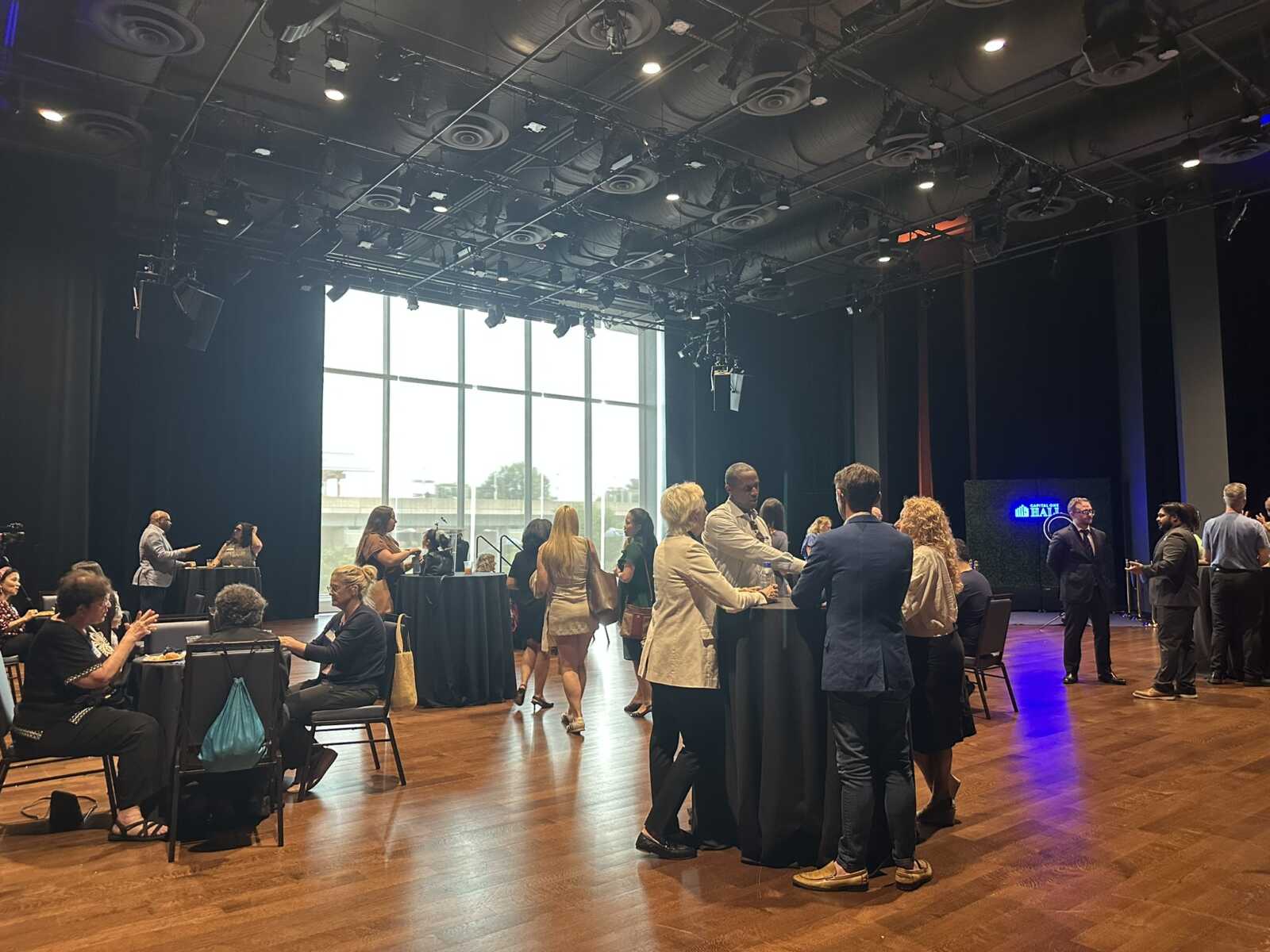 Professionals mingle at a reception in a large event space with wooden floors, high ceiling, and large windows in the background.