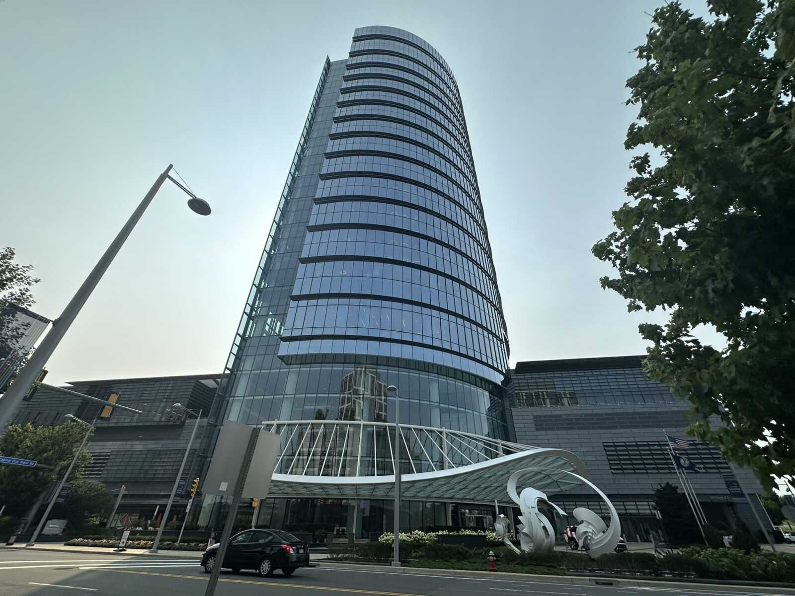 Tall curved glass office tower dominates the street, with a glass-covered plaza and sculptural installation at the base; trees on the right and a street lamp in the foreground.