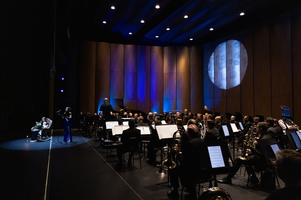 Capital Wind Symphony performs at Capital One Hall (courtesy Capital One Hall) | FFXnow Orchestra performing on stage with a conductor and a soloist in a blue dress under blue stage lighting in a concert hall.