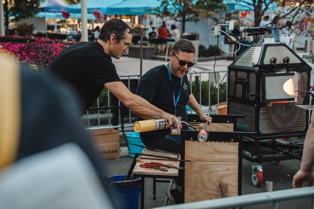 Two men work at an outdoor glassmaking station, one heating a glowing glass piece with a torch while the other assists nearby.