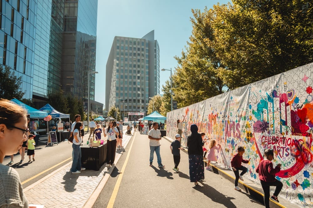 City street festival with tents, people strolling, and a colorful graffiti wall behind them at a sunny downtown event.