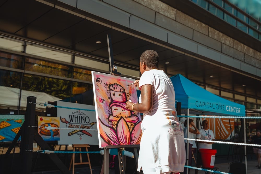 Artist painting a pink-orange portrait on a canvas outdoors, with event banners and a blue Capital One Center tent nearby.