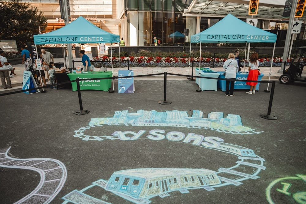 Outdoor street festival scene with two blue 'Capital One Center' tents, tables of volunteers, and attendees browsing booths amid a flower bed backdrop; chalk art on the pavement features a circular cityscape and tram.