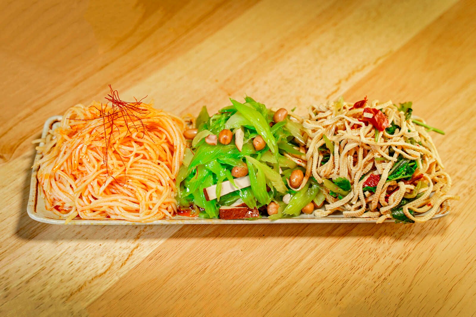 Tri-color noodle platter with three sections: spicy orange noodles on the left, green vegetable and peanut mix in the center, and crispy noodle salad on the right.