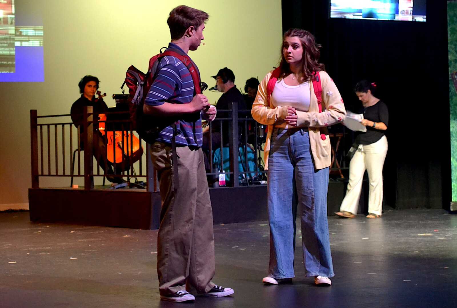 Two teens stand on a stage facing each other, backpacks on