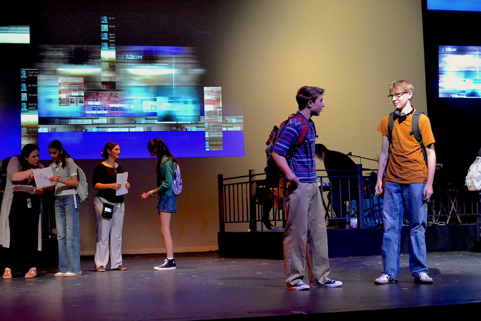 Group of students on a stage, some holding papers and backpacks, chatting while a blue digital display projects in the background.