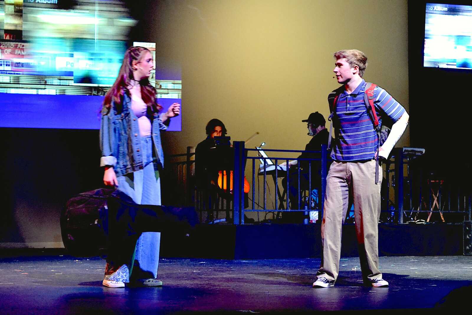 Two teens perform on stage: girl in a denim jacket facing a boy with a striped polo and backpack, with musicians in the background.
