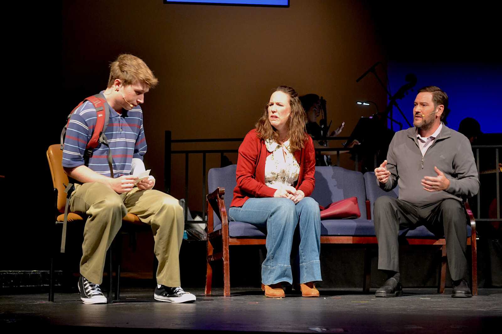 Three people sit on stage: a young man in a striped polo with backpack on the left, a woman in a red cardigan in the middle, and a man in a gray sweater speaking on the right.