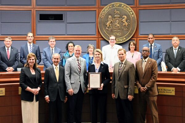 Group of officials posing in a formal chamber; woman at center holds an award plaque in front of a Fairfax County seal panel.