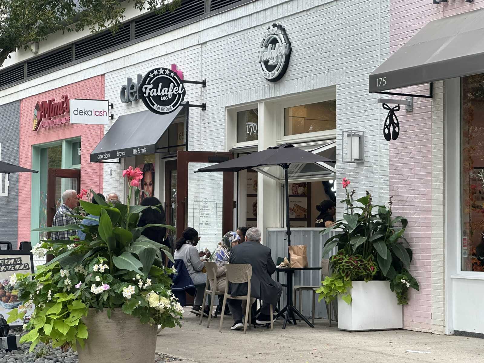 Outdoor seating on a sidewalk in front of colorful storefronts, with people dining under umbrellas and large flower planters nearby.