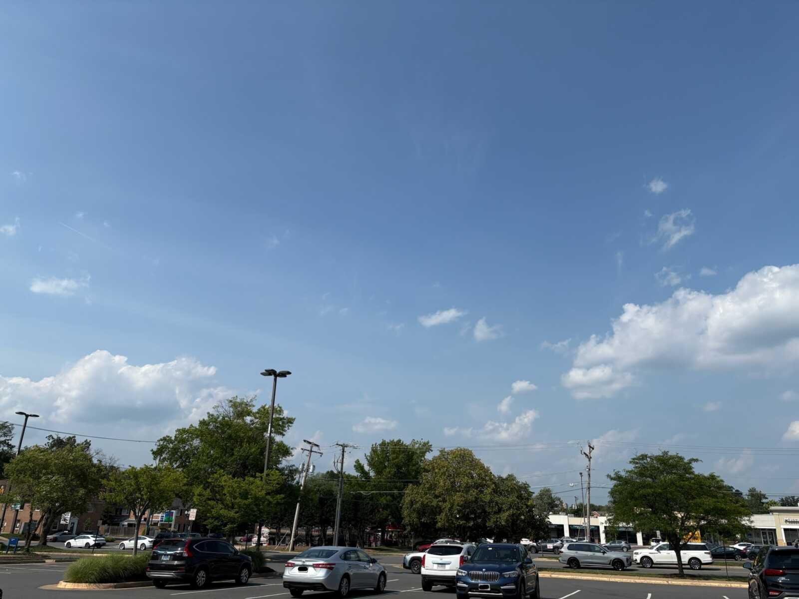 Parking lot with several parked cars, trees along the edge, and streetlights under a clear blue sky with a few clouds.