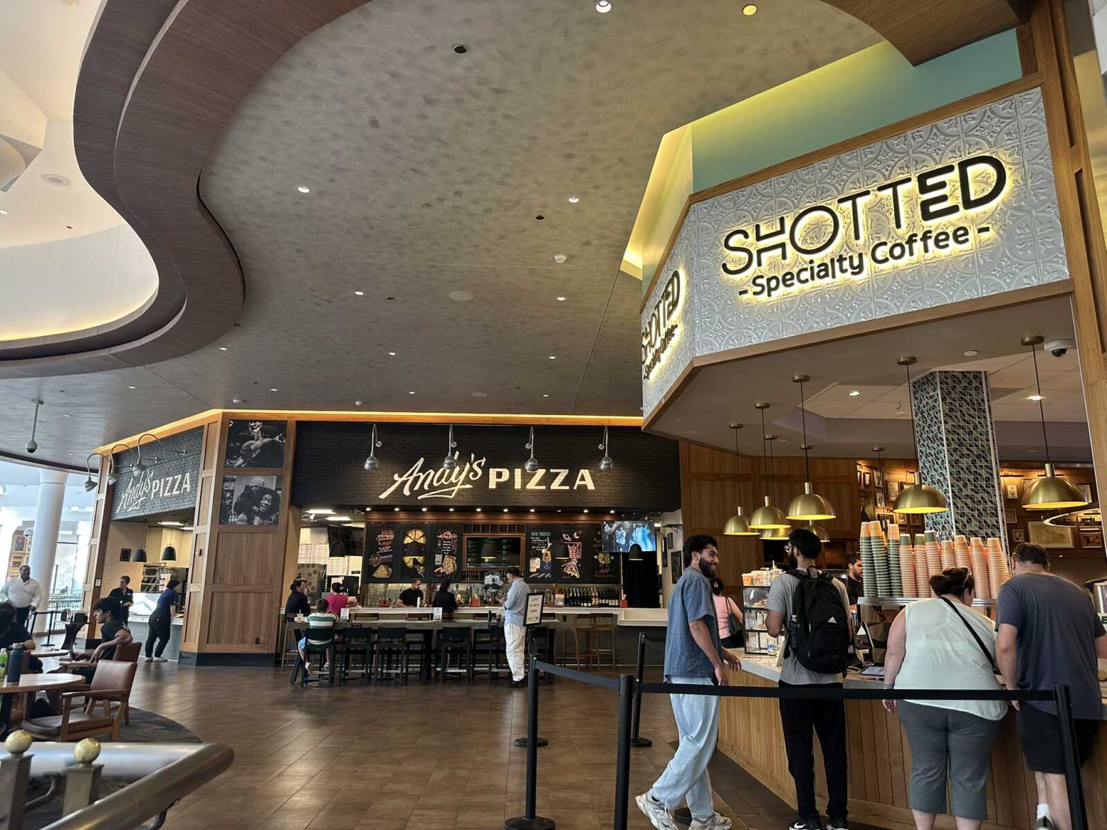 Inside a modern food court with Anay's Pizza and SHOTTED Specialty Coffee storefronts, customers in line and seated nearby.