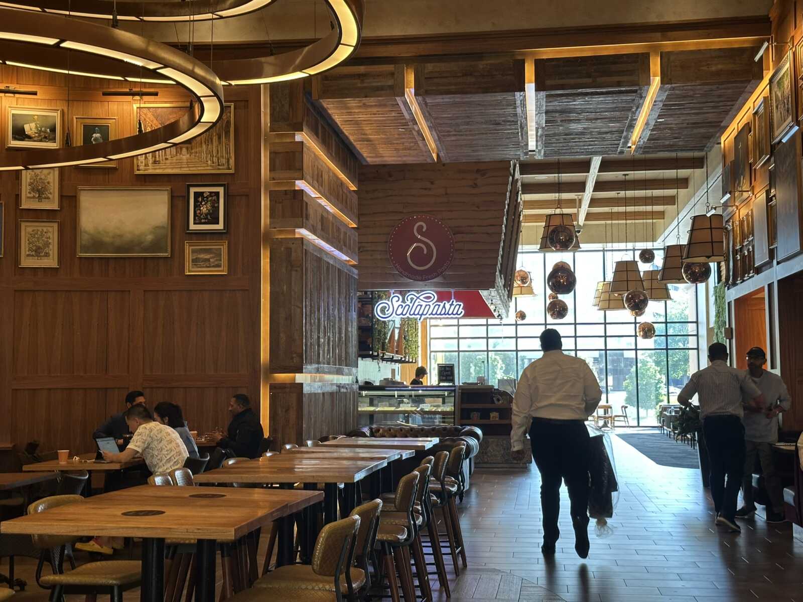 Cozy restaurant interior with wood-paneled walls, framed art, and warm circular pendant lights; customers sit at tables on the left.