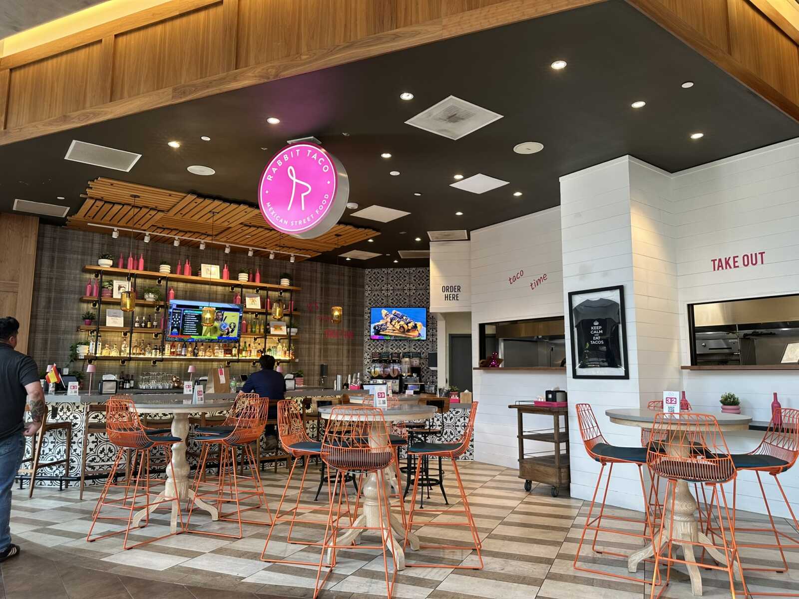 Interior of a modern taco shop with orange wire chairs, a long bar, and pink circular Rabbit Taco sign overhead.
