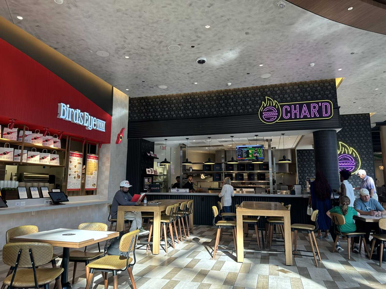 Food court scene with two restaurant counters: Bird's Eye Thai on the left and CH'ARD on the right, and diners at wooden tables in the foreground.