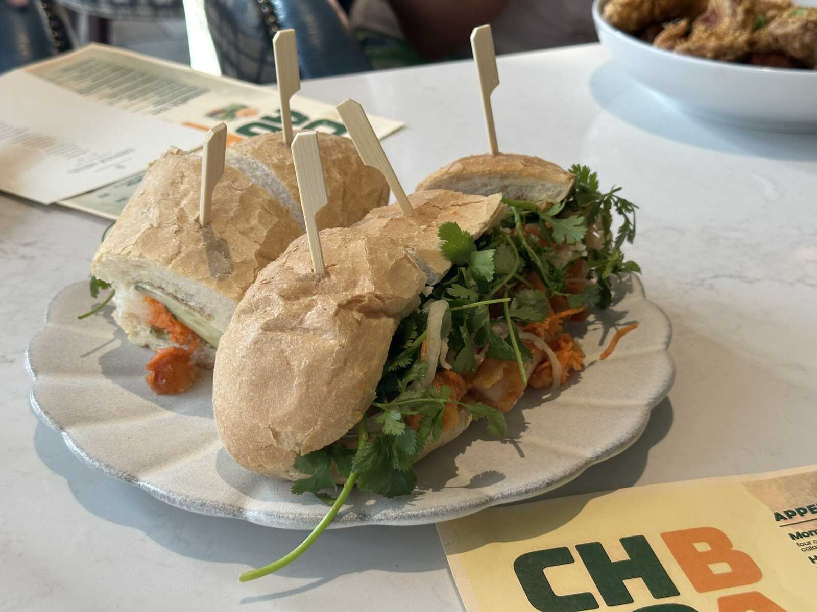 Banh mi-style sandwich with cilantro, carrot, and vegetables on a crusty baguette, served on a plate with skewers.