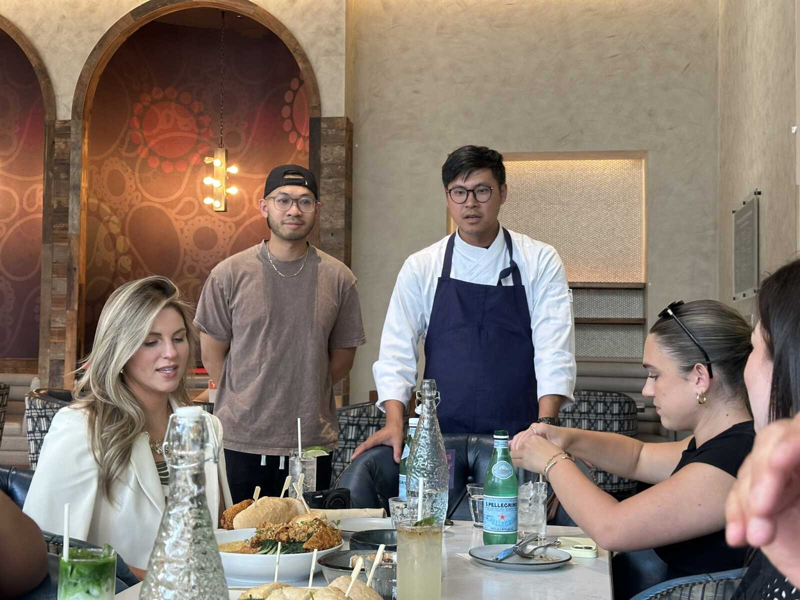Waiter in white shirt and navy apron speaks to diners at a restaurant table with food and drinks in front of them.