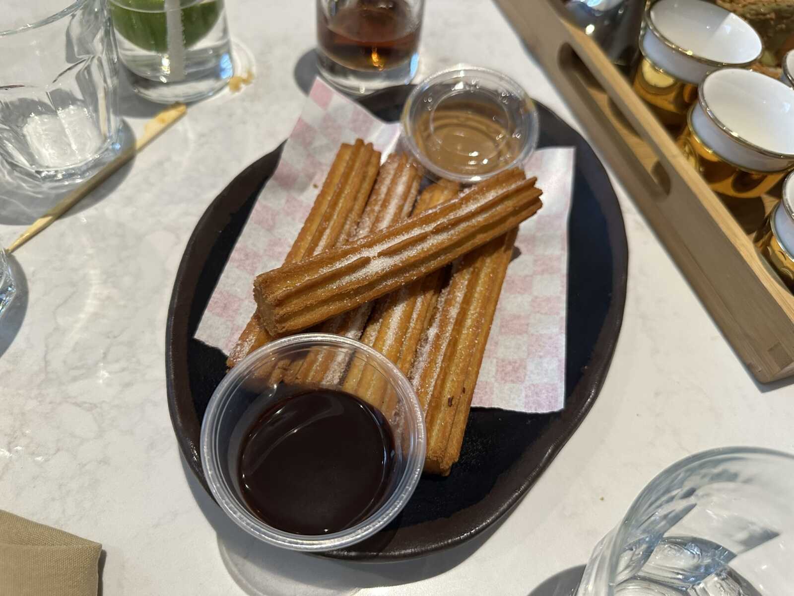 Churros dusted with sugar served in a black tray with dipping sauces (chocolate and caramel) on a checkered napkin.