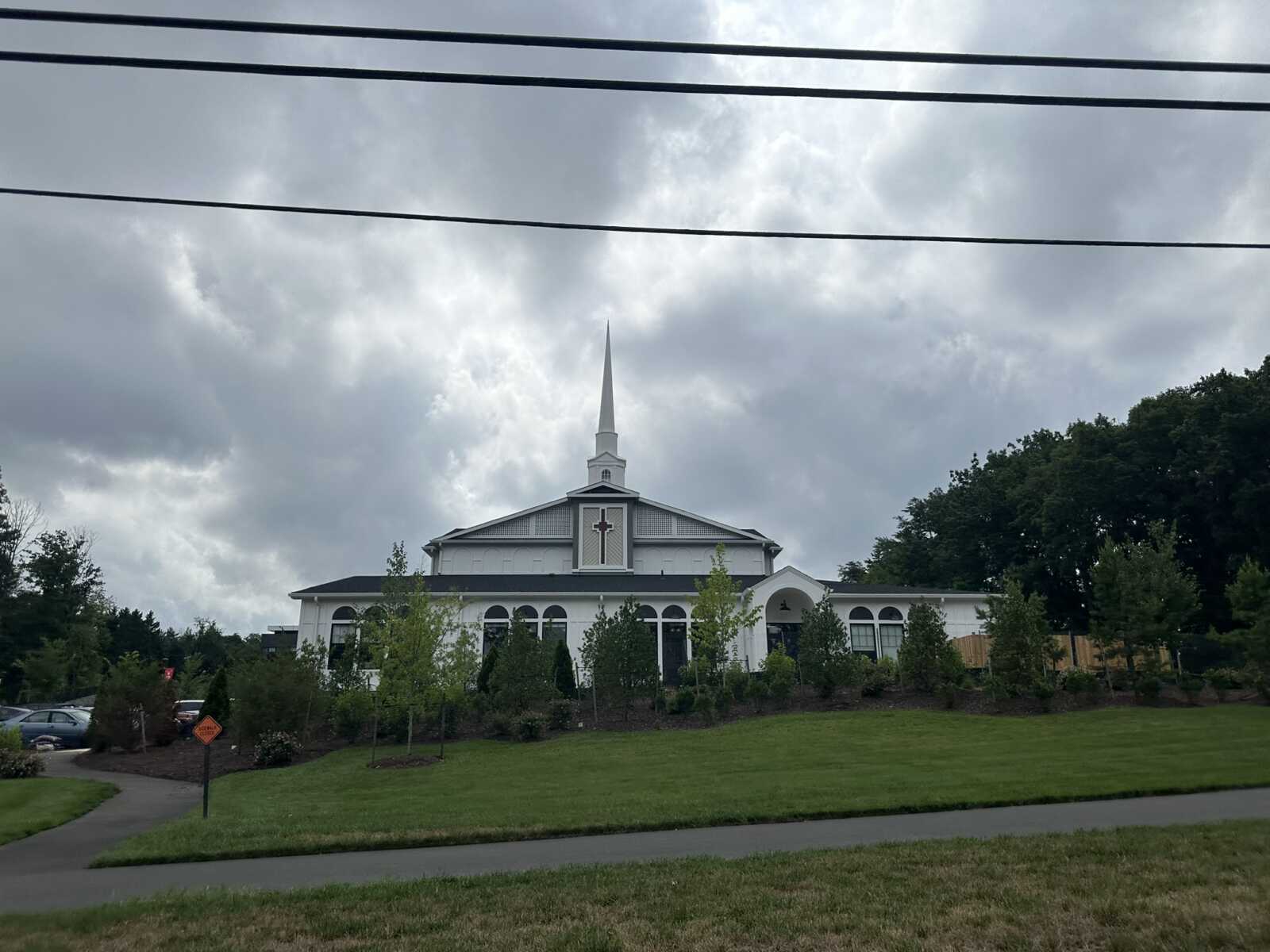 White church with a tall steeple and cross, arched windows, set in a landscaped lawn with trees under a cloudy sky