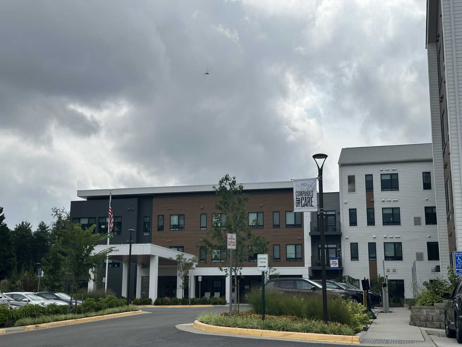 Modern mixed-use buildings with brown and gray facades under a cloudy sky; an American flag on a pole near the entrance and a banner on a lamp post reading CAREs/CARE.