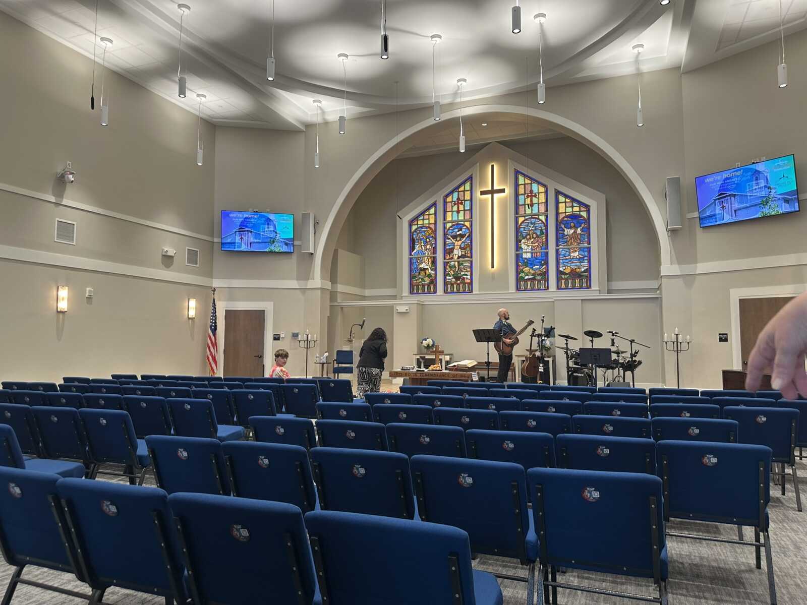 Church sanctuary with rows of blue chairs facing a worship stage, where a musician plays guitar beneath a large stained-glass backdrop and cross.