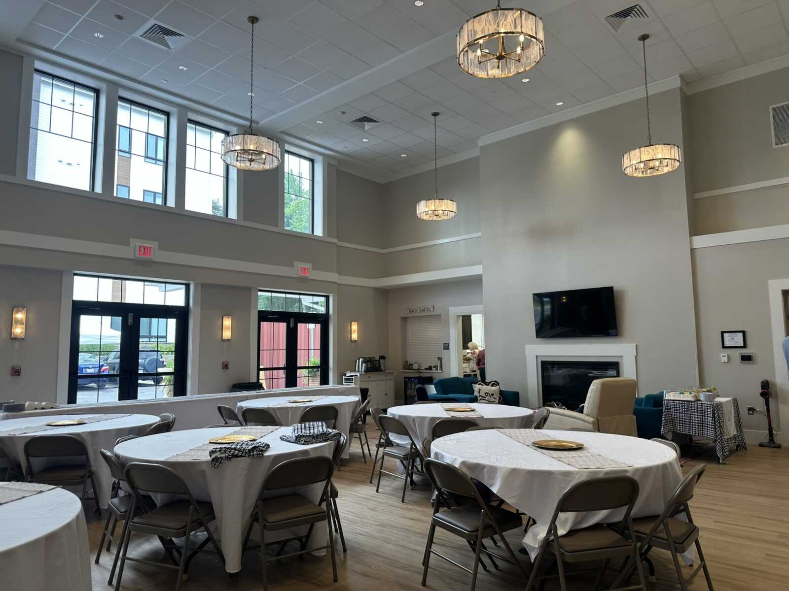 Spacious banquet hall with round tables covered in white cloths, metal folding chairs, and chandelier ceiling lights.