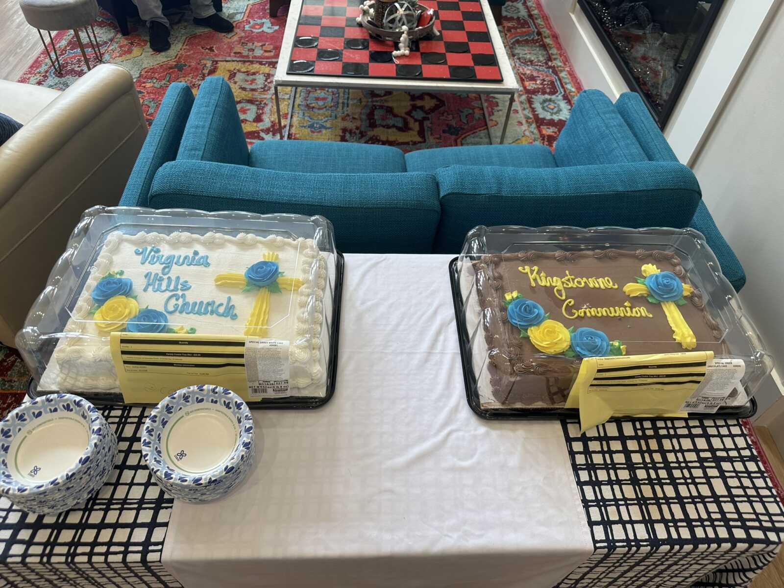 Two rectangular celebration cakes with blue and yellow sugar flowers on a table in a cozy living room setting with blue chairs and a colorful rug.