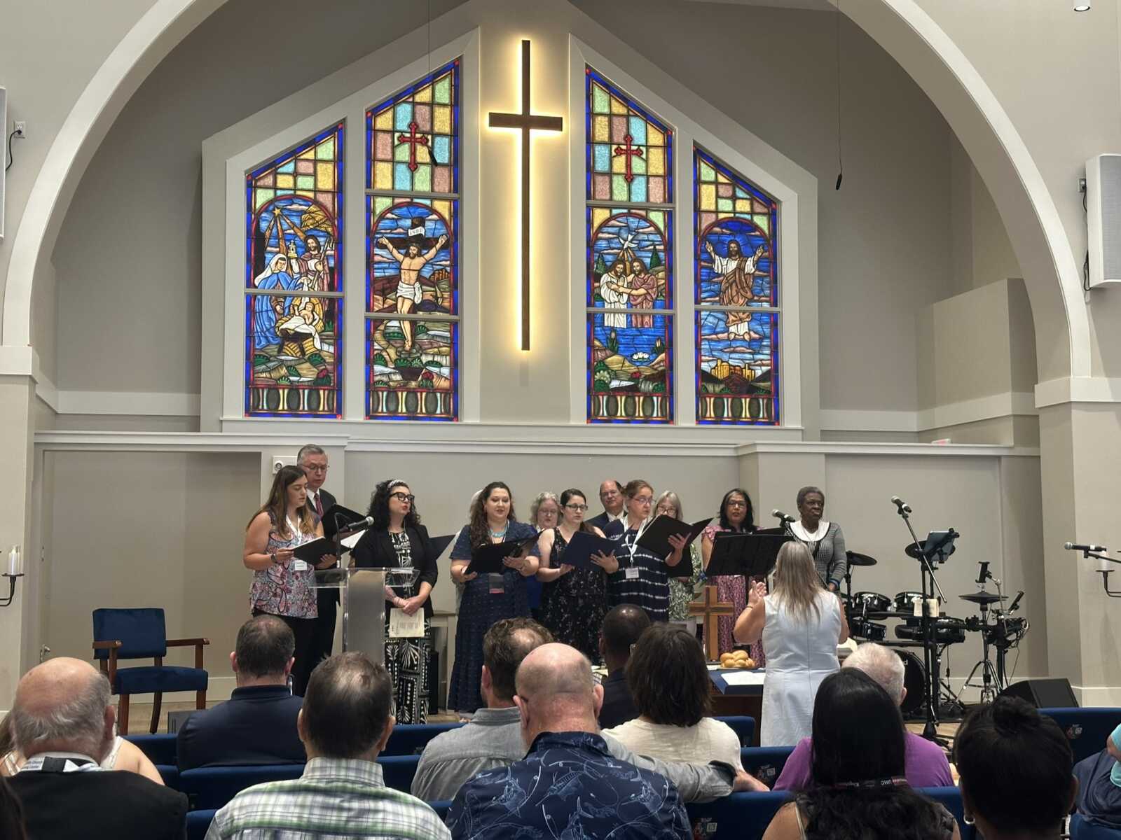 Chorus of diverse singers performing on a church stage with stained-glass windows and a glowing cross behind them.