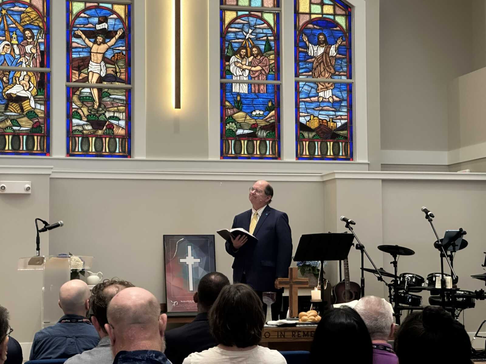 A man in a dark suit and yellow tie stands at a pulpit reading from a book during a church service, with colorful stained-glass windows behind him and microphones on stands nearby.