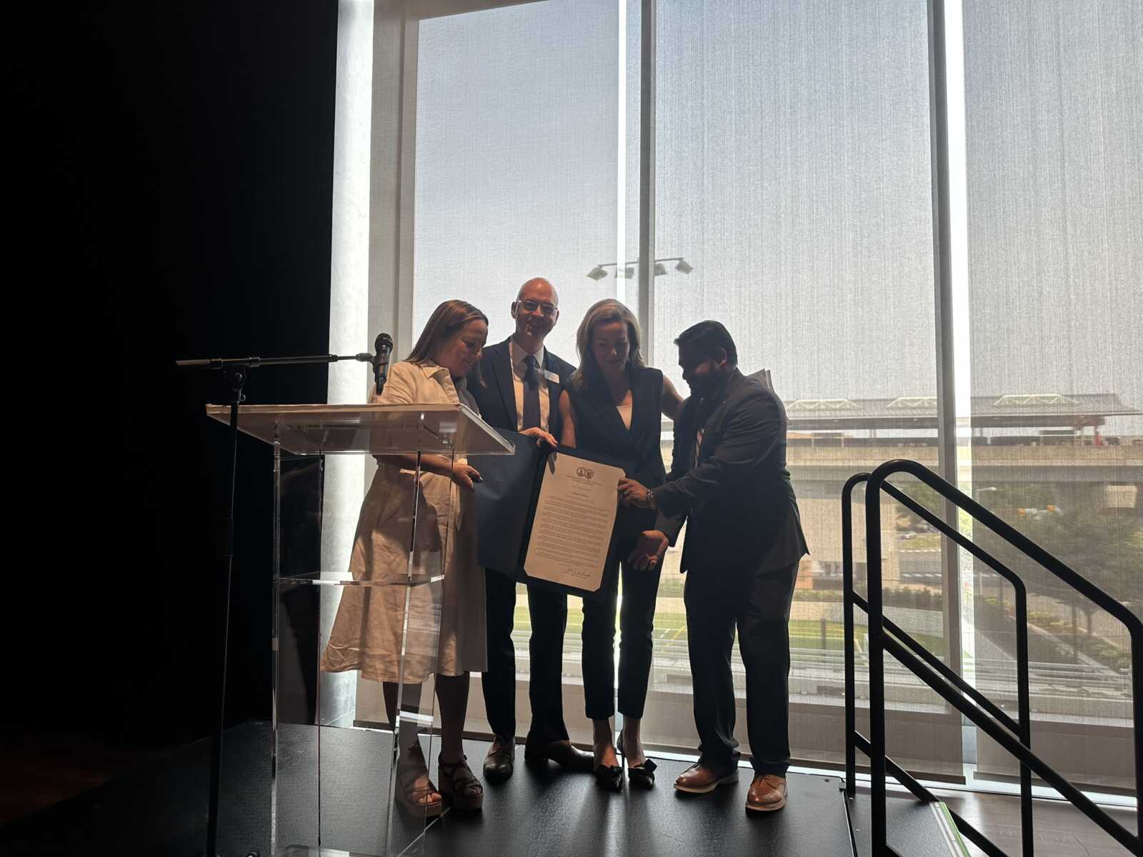 Four professionals in business attire gather on a stage, examining a large document during a ceremony with a podium and microphone nearby.