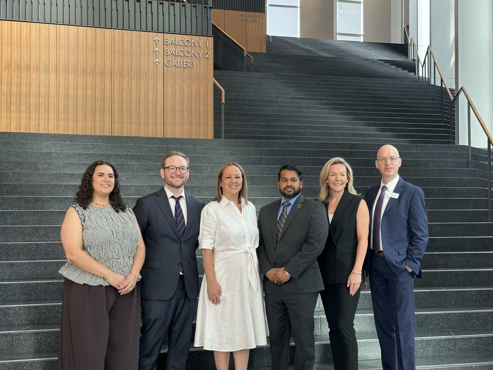 Six professionals in business attire pose on a wide indoor stairway, smiling for a group photo on a dark stone staircase.