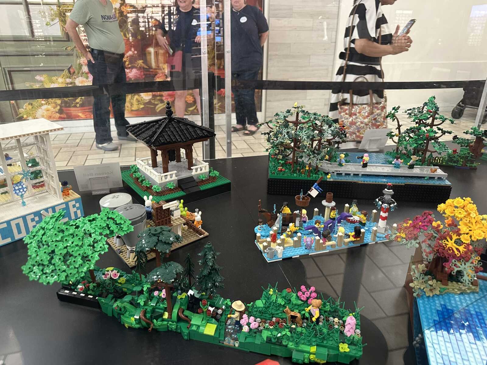 LEGO diorama display with a black-roofed gazebo in a garden, surrounded by trees, flowers, and small figurines in a storefront window