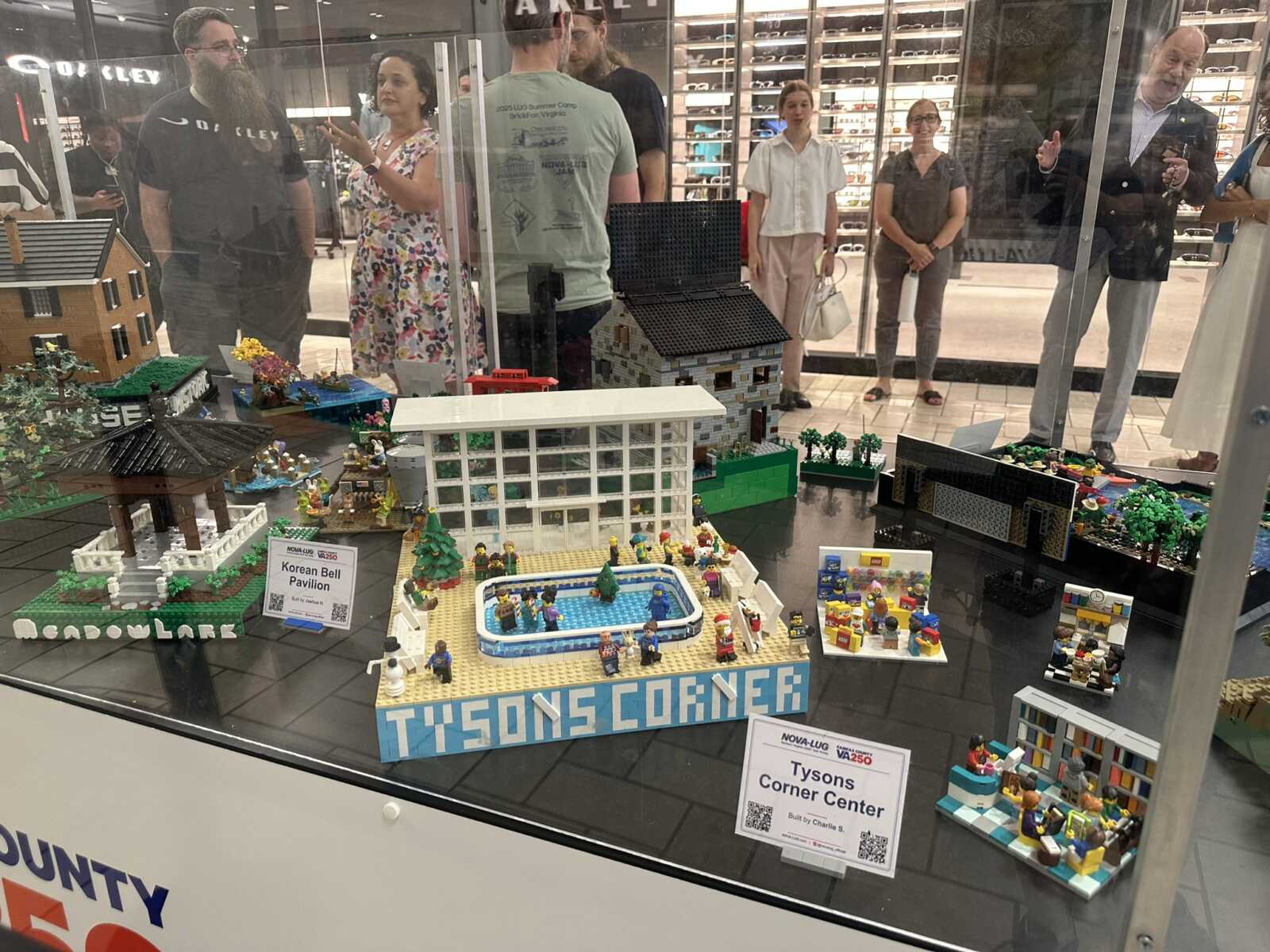 Lego city display titled 'Tysons Corner' with multiple buildings, trees, and minifigures visible through a storefront glass window.