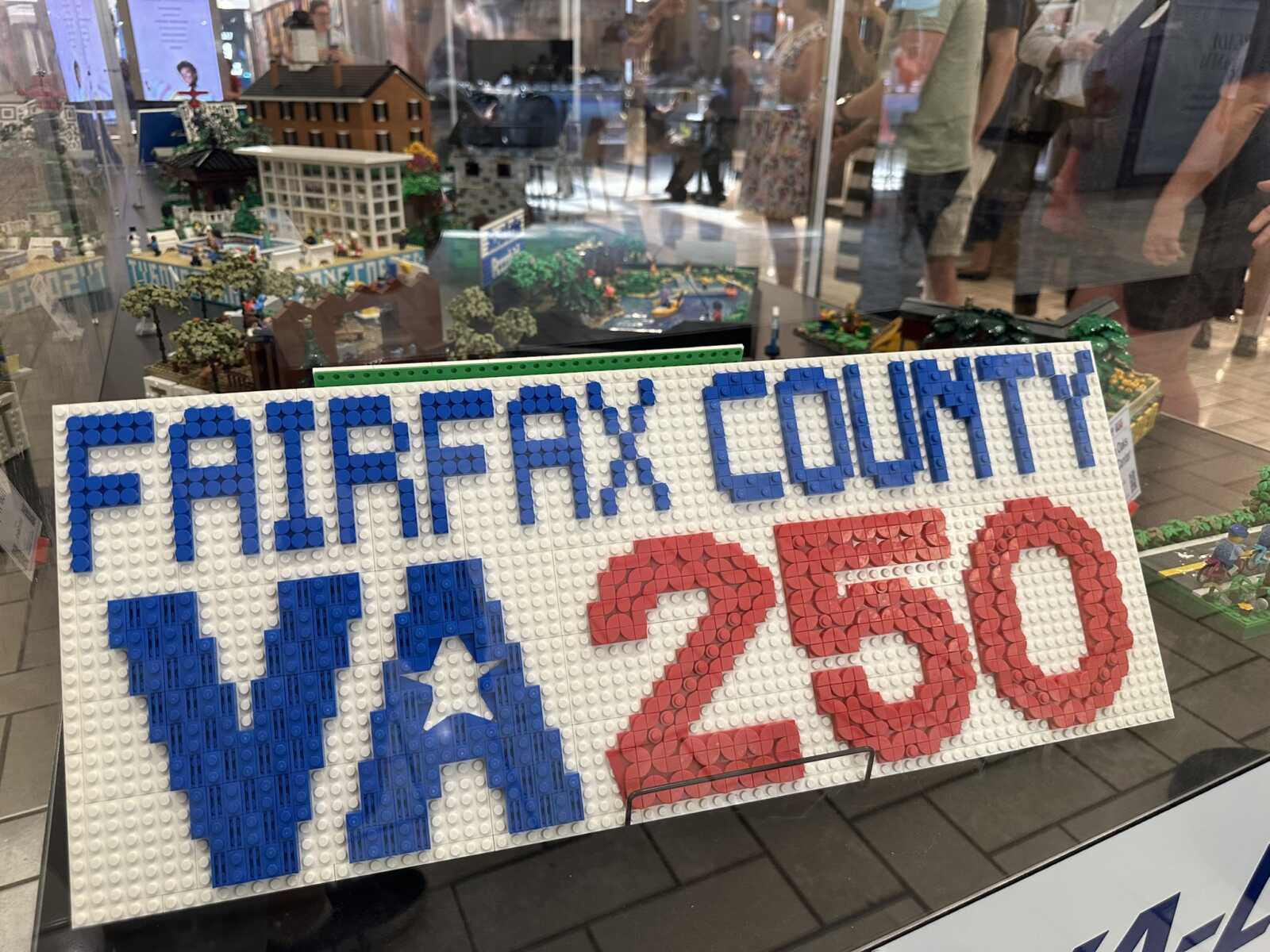 Close-up of a LEGO display sign in a shop window, with blue letters on white bricks and red numbers, beside a miniature city scene behind the glass.