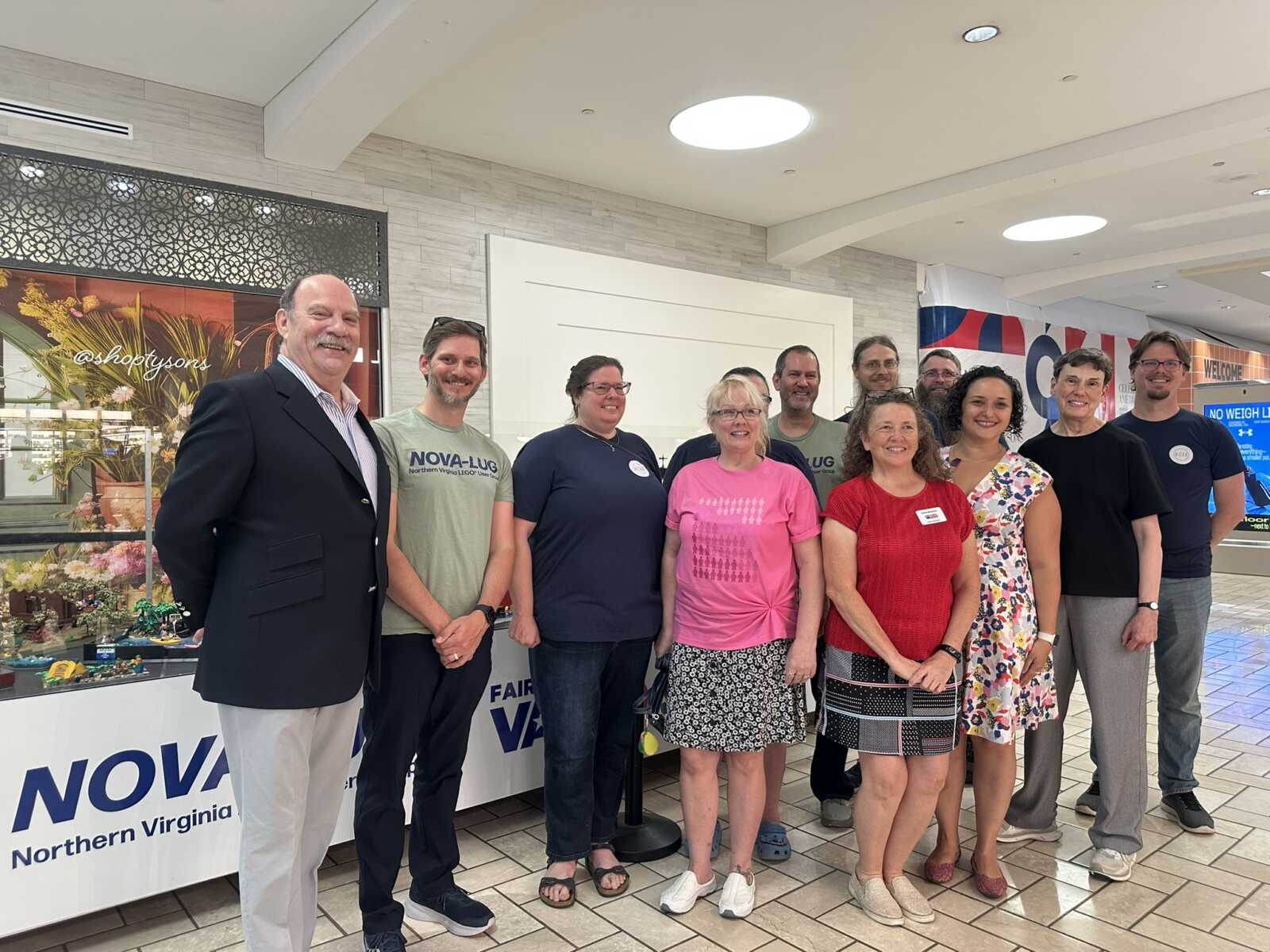 Group of twelve adults posing together and smiling at a community event inside a shopping center near a NOVA banner and flower display.
