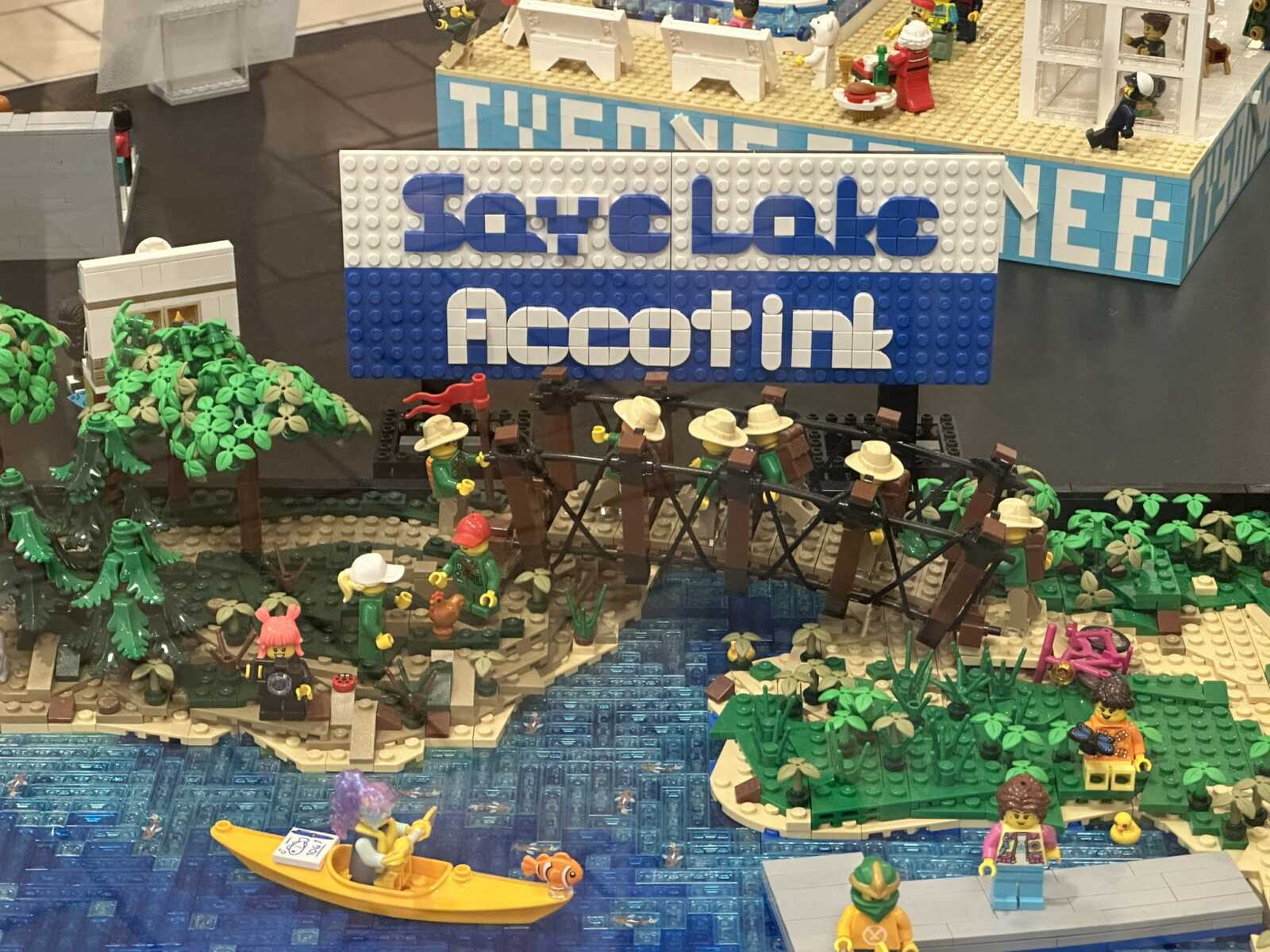 LEGO diorama with a large blue-and-white Facebook sign, a wooden bridge over a blue river, and minifigures in a jungle setting nearby with trees and plants.