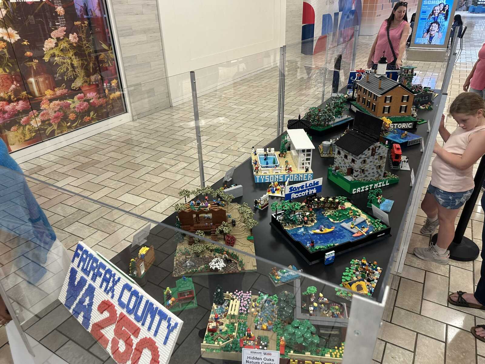 LEGO city diorama display in a mall window, with multiple buildings and a girl watching nearby.